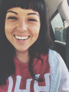 Natalie, wearing a red shirt with a white sweater sitting in her car with an enormous smile. 