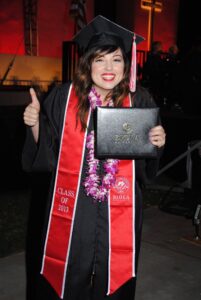 Natalie at her college graduation holding a diploma and wearing a big smile. Natalie is wearing a black gown with a red ribbon that reads "class of 2013."