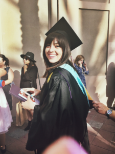 Natalie at her graduation for her master's degree in a black cap and gown with a blue hood. Big smile, diploma in hand. 