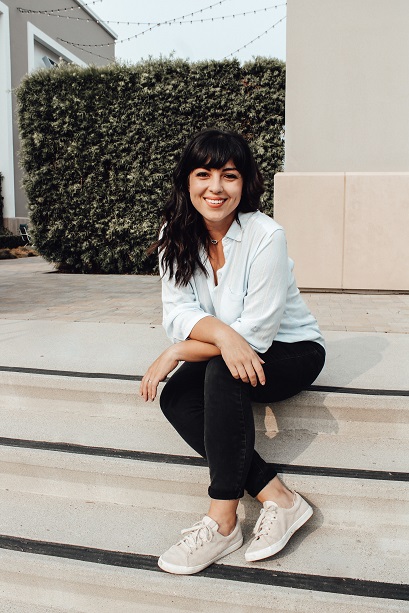 Natalie sitting outdoors on some stairs. Arms crossed, smiling. Natalie is wearing a blue shirt, black pants, and pink shoes.